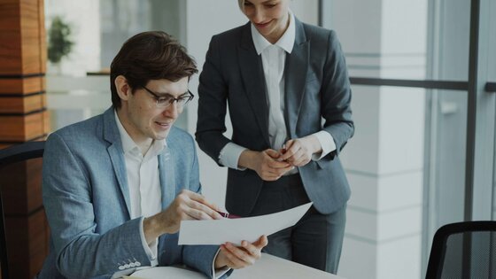 Eine Frau und ein Mann diskutieren ein Dokument. Der Mann sitzt am Tisch, die Frau steht neben ihm. Beide befinden sich in einem Bürogebäude.